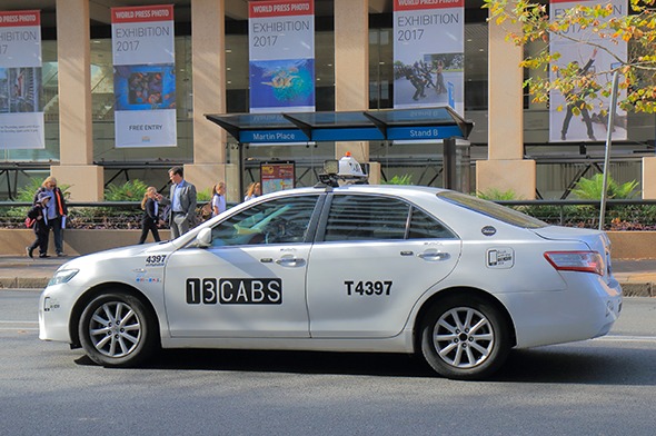A photograph of a grey taxi on a Sydney city street during the day. "13 CABS" is written on the passenger door