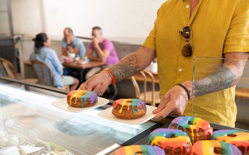 Someone with tattoos collecting two rainbow cronuts from the counter at Infinity Bakery in Darlinghurst