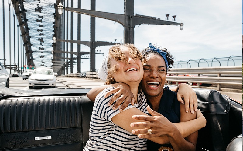 Two women hugging in the back of a convertible car travelling over the Harbour Bridge